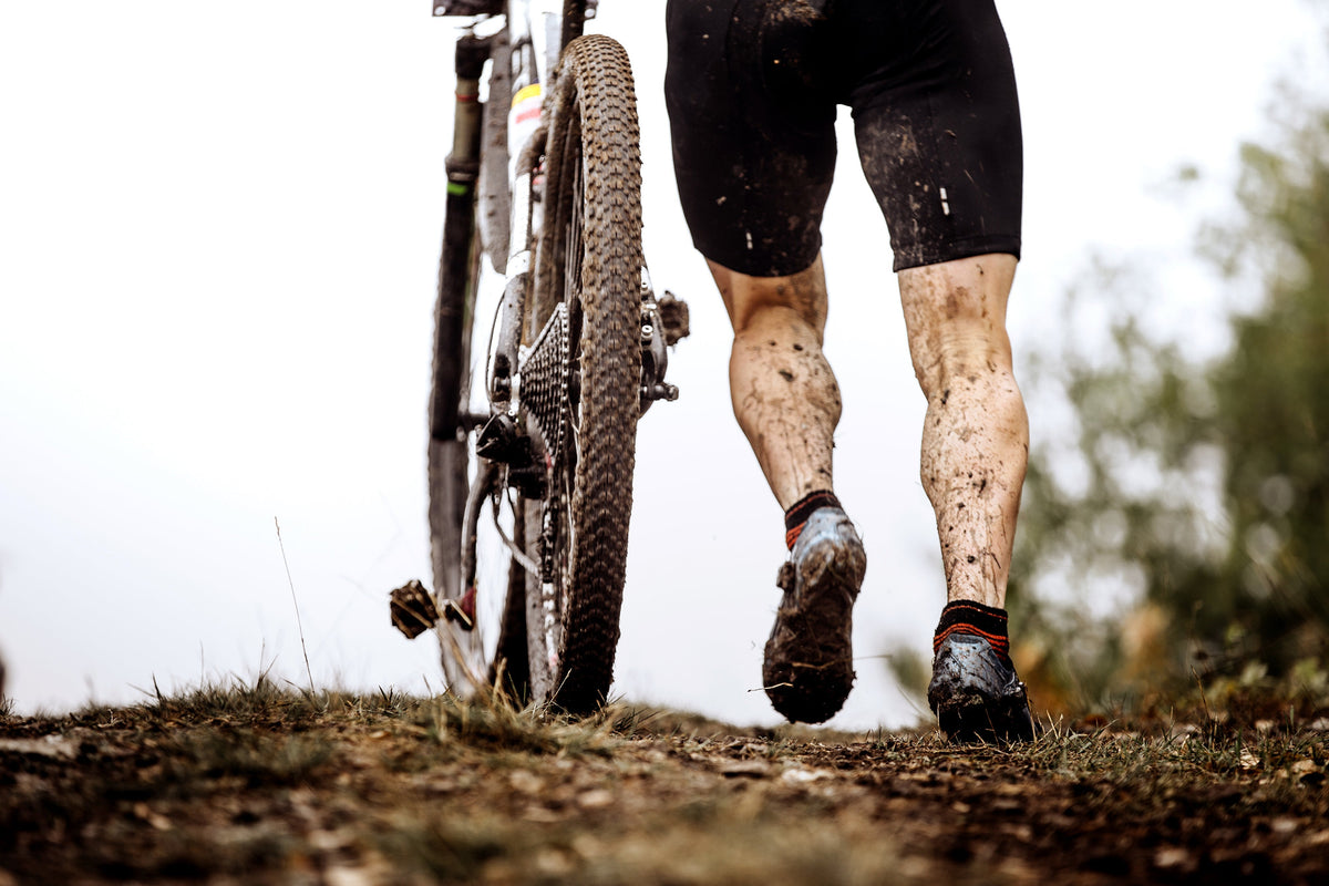 Rider with mud-covered legs carrying a dirty bike up a muddy slope
